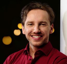 Ein Mann in rotem Hemd lächelt in die Kamera. Unscharfer Hintergrund mit Lichtern.