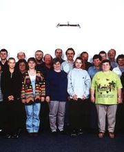 Gruppe von etwa 30 Personen steht in zwei Reihen in einem Raum und lächelt in die Kamera.