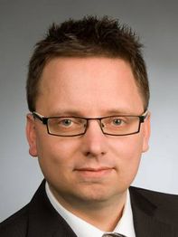 Ein Mann mit Brille und dunklem Haar im Anzug, schaut direkt in die Kamera vor grauem Hintergrund.