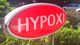 Rotes Schild mit der Aufschrift "HYPOXI" vor einer grünen Hecke und mehreren geparkten Autos.