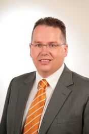 Ein Mann im Anzug mit orange-gestreifter Krawatte lächelt vor neutralem Hintergrund.