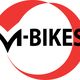 Logo met tekst "M-BIKES" in zwart binnen een rode cirkel.