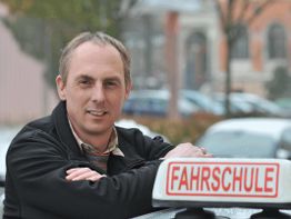 Ein Mann lehnt an einem Auto mit Fahrschule-Schild. Hintergrund mit unscharfen Gebäuden.
