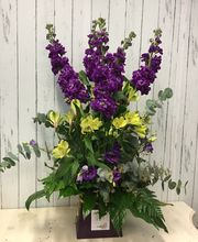 Ramo de flores púrpuras y amarillas con hojas verdes sobre fondo de madera blanca.