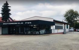 Autohandel mit moderner Glasfront, umgeben von Bäumen und einem gepflasterten Parkplatz.