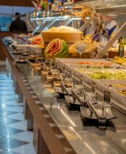 Buffet variado com pratos diversos e frutas decorativas, em salão com piso xadrez.