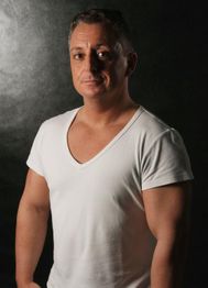 Mann in weißem T-Shirt vor dunklem Hintergrund, schaut neutral in die Kamera.