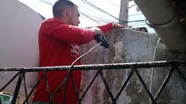 Homem com camisa vermelha usa uma furadeira elétrica em uma parede ao ar livre.