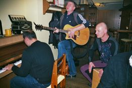 Drei Männer musizieren: einer spielt Gitarre, einer Klavier, der dritte schlägt auf eine Cajón.