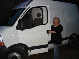 Frau neben einem weißen Lieferwagen mit geöffneter Fahrertür, im Freien aufgenommen.