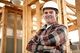 Smiling construction worker in a hard hat and plaid shirt standing in a wooden building frame.