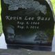 Black gravestone with "Kevin Lee Bass" and dates, sky with clouds, and tree engraving.