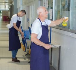 Two janitors wearing aprons clean windows and stack chairs in a hallway.