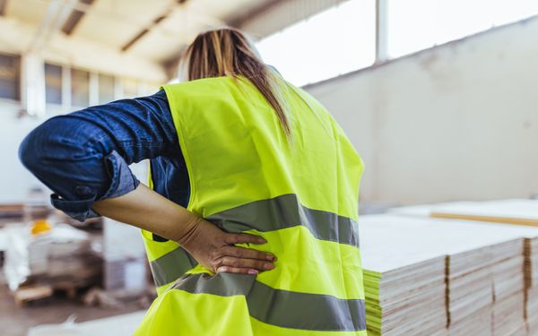 Eine Lageristin hält sich ihren Rücken mit der linken Hand. Von der körperlichen Arbeit hat sie Rückenschmerzen.