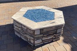Hexagonal stone fire pit with blue glass center, on a paved patio.