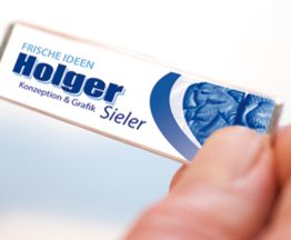 Hand hält einen kleinen rechteckigen Gegenstand mit blauem Text: "Frische Ideen, Konzeption & Grafik".