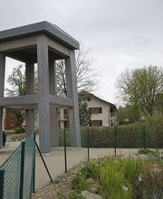 Grau-betonierter Pavillon neben einem Weg, umgeben von Häusern und Bäumen an einem bewölkten Tag.