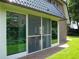 Hausfassade mit großen Glas-Schiebetüren, umgeben von grünem Rasen und Bäumen.