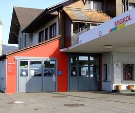 Tankstelle mit MIGROL-Logo, rote und weiße Gebäude, blauer Himmel im Hintergrund.