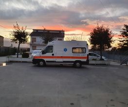 Ambulanza parcheggiata in una piazza al tramonto con cielo nuvoloso e alberi sullo sfondo.