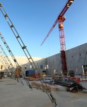 Baustelle mit Betonmauern und rotem Kran bei klarem Himmel. Gerüste und Baumaterialien sichtbar.
