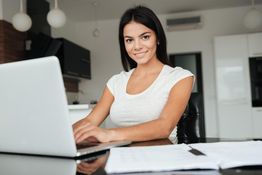 Woman smiling, typing on a laptop at a desk with papers in a modern room.