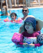Criança com bóias e chapéu brinca na piscina, acompanhada por outras pessoas ao fundo.