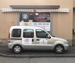 Silbernes Taxi parkt vor einem Schaufenster mit "Best Taxi Lugano" und einer Telefonnummer.