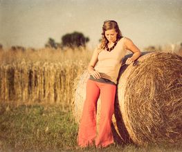 Frau lehnt an Heuballen auf Feld, trägt rote Hose, blondes Haar, friedliche Landschaft.