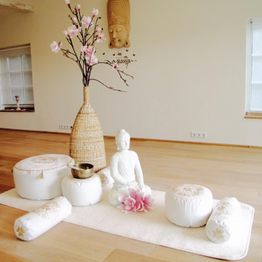 Meditationsraum mit Buddha-Statue, Kissen, Blüten und Zweig in einer Vase auf hellem Holzfußboden.