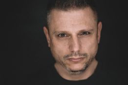Ein Mann mit kurzem Haar schaut ernst in die Kamera, dunkler Hintergrund.