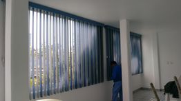 Hombre instala persianas verticales azules en una sala luminosa con paredes blancas.