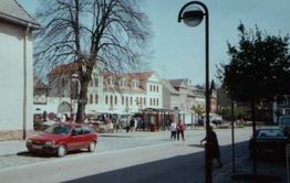 Straße mit parkendem rotem Auto, Menschen spazieren, historische Gebäude und Bäume im Hintergrund.