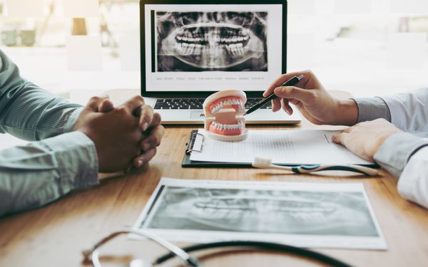 Gespräch zwischen Patient und Kieferorthopäde. Der Kieferorthopäde deutet mit einem Kugelschreiber auf ein Model eines Gebisses. Im Hintergrund steht ein Laptop auf dem ein Röntgenbild eines Kiefers zu sehen ist.