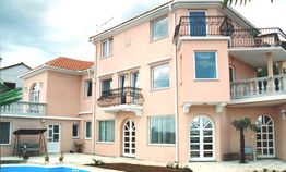Großes, rosa Haus mit Balkon, gewölbten Fenstern und Pool im Garten bei bewölktem Himmel.