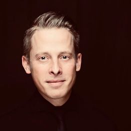 Ein Mann in schwarzem Hemd vor dunklem Hintergrund, schaut direkt in die Kamera.