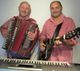 Zwei Männer in karierten Hemden spielen Akkordeon und Gitarre, ein Keyboard steht vor ihnen.