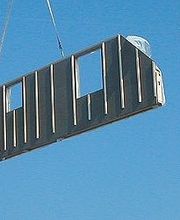 Kran hebt Wandsegment auf Baustelle in die Luft, blauer Himmel im Hintergrund.