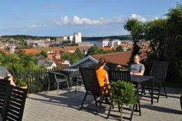 To personer sidder ved et bord på en terrasse med udsigt over en by og havet i solskinsvejr.