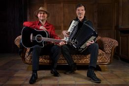 Zwei Männer auf einem Sofa, einer mit Gitarre, der andere mit Akkordeon, vor Holzwand.