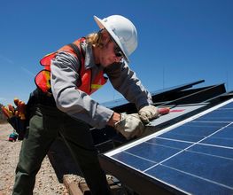 Persona instalando panel solar con casco y chaleco, cielo despejado de fondo.