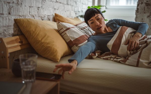 Erschöpfte Frau liegt im Bett und greift nach einem Glas Wasser auf dem Nachttisch.