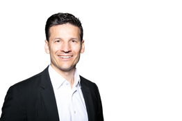 Eine lächelnde Person in Anzug mit weißem Hemd vor schwarzem Hintergrund.