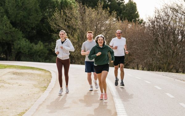 Gruppe von vier Personen unterschiedlichen Alters beim gemeinsamen Joggen auf einer Straße im Grünen.