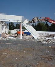 Ein Bagger reißt ein Gebäude bei sonnigem Wetter ab. Im Hintergrund sind Bäume zu sehen.