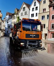 Straßenreinigungsfahrzeug sprüht Wasser in einer Kopfsteinpflasterstraße vor bunten Gebäuden.
