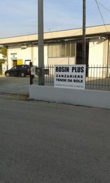 Cartello "ROSIN PLUS" su recinzione, ingresso aziendale con zanzariere e tende da sole.