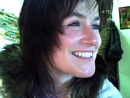 Eine Frau mit braunen Haaren lächelt zur Seite, im Hintergrund sind Bücher sichtbar.