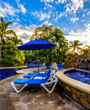 Piscina con tumbonas azules, sombrillas y palmeras bajo un cielo despejado.