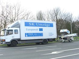 LKW der Firma SK Umzüge mit Aufzuganhänger auf einer Straße, Bäume im Hintergrund.